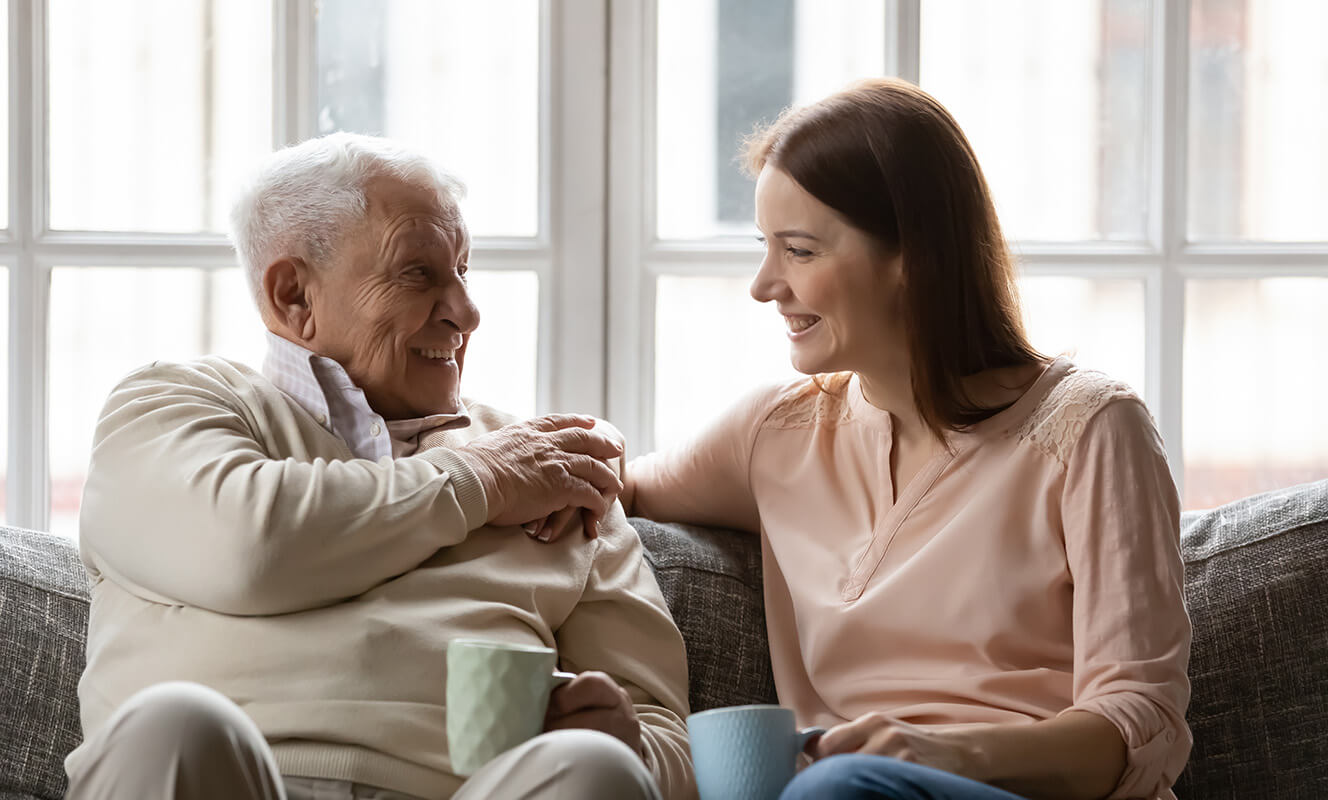 Elderly man and young woman smiling while sitting on a sofa, holding mugs